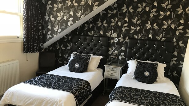 A bedroom with two beds, a black upholstered headboard, a bedside table with a lamp, and a window with curtains. - Canterbury Lodge (Canterbury)