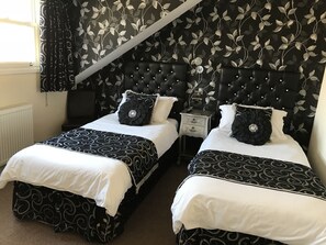 A bedroom with two beds, a black upholstered headboard, a bedside table with a lamp, and a window with curtains. - Canterbury Lodge (Canterbury)
