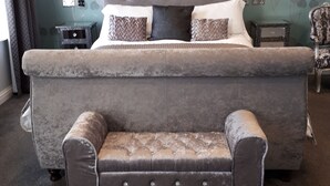 A bedroom with a large bed, a bedside table, a chair, and a wall with floral wallpaper. - Canterbury Lodge (Canterbury)