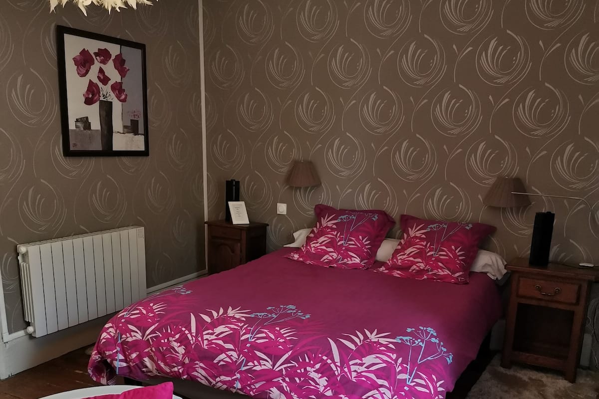 A bedroom with a bed, two nightstands, a lamp, a radiator, and two white chairs with pink cushions.
