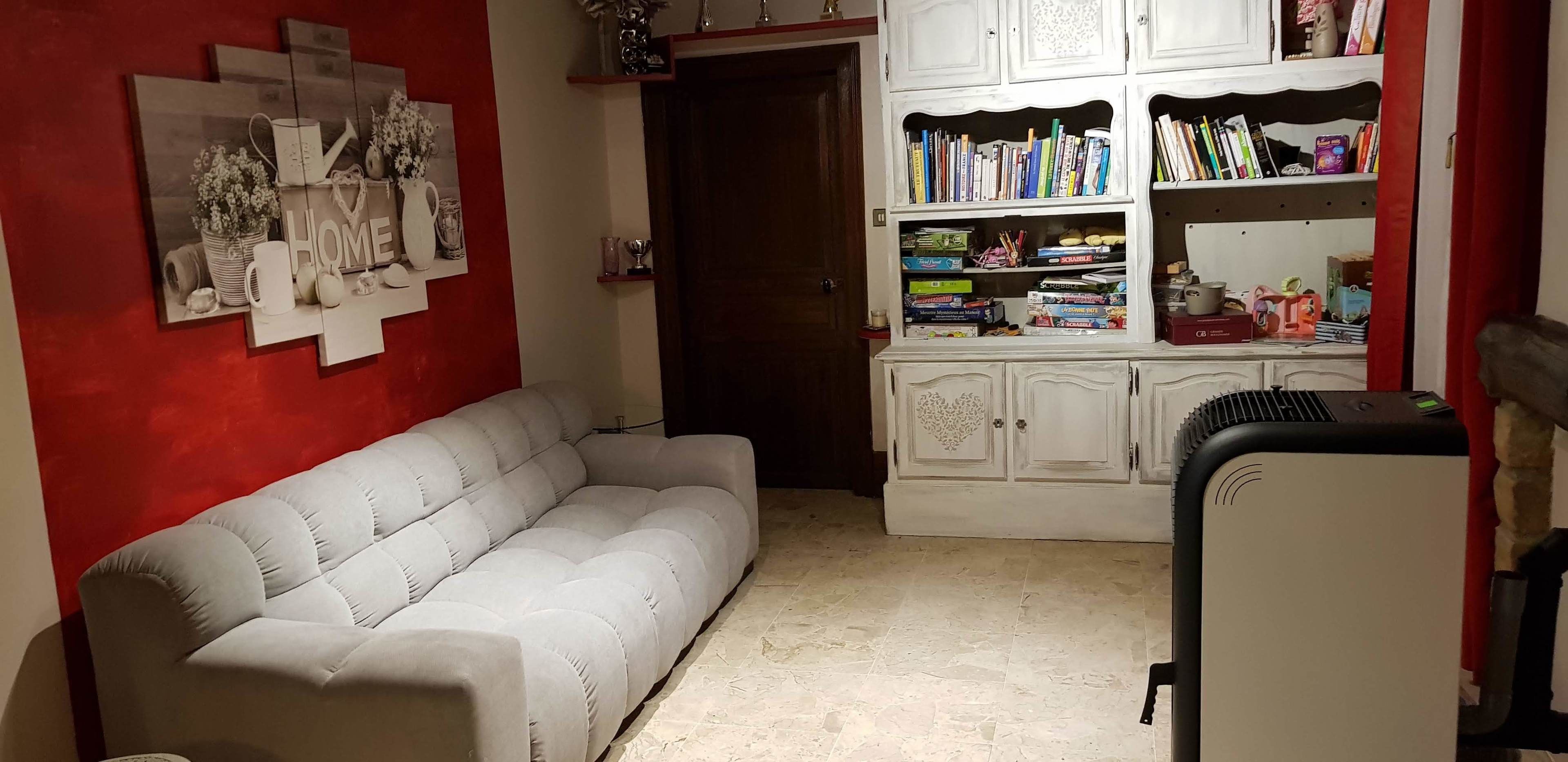 A living room with a grey sofa, a white cabinet with books and decorative items, and a red wall with framed pictures.