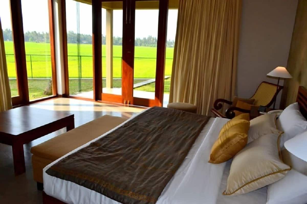 Quarto de hotel com uma cama grande, um banco de madeira, uma cadeira e vista para um campo verde através de grandes janelas.
