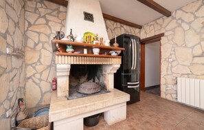 A stone-walled room with a fireplace, a wooden shelf with various items, a black refrigerator, and a tiled floor.