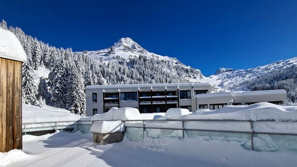 Un bâtiment moderne à plusieurs étages, entouré de montagnes et d’arbres couverts de neige.
