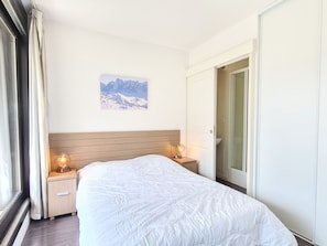 Quarto de hotel com uma cama grande, cabeceira de madeira, duas mesas de cabeceira com abajures, um quadro na parede e uma porta deslizante para o banheiro.