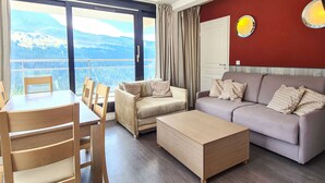 Un salon moderne comprenant un canapé, une table basse et un coin repas avec des chaises. Une grande fenêtre offre une vue panoramique sur les montagnes et un lac.
