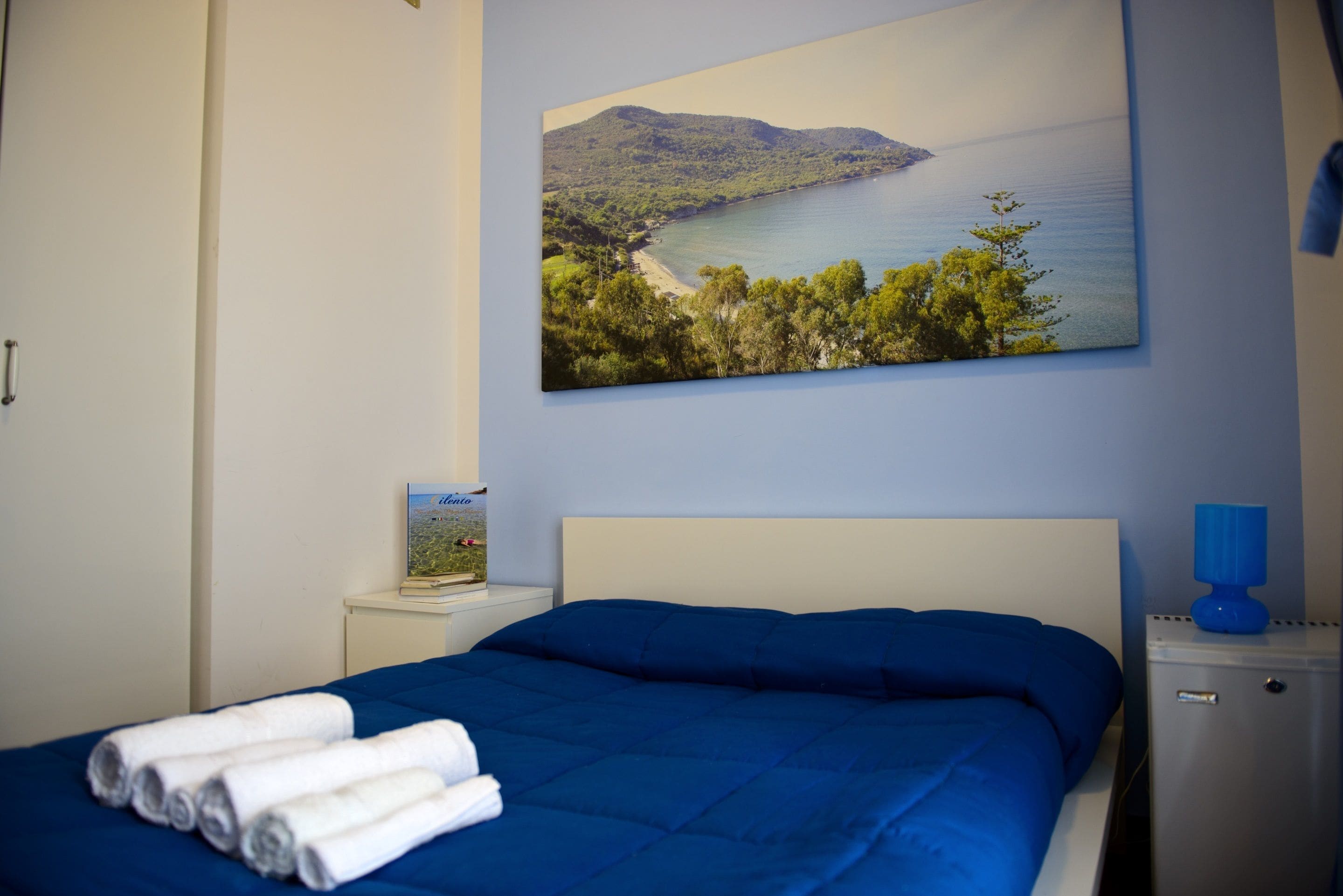 A bedroom with a bed, a painting of a coastal landscape, a bedside table with a lamp, and a small cabinet.