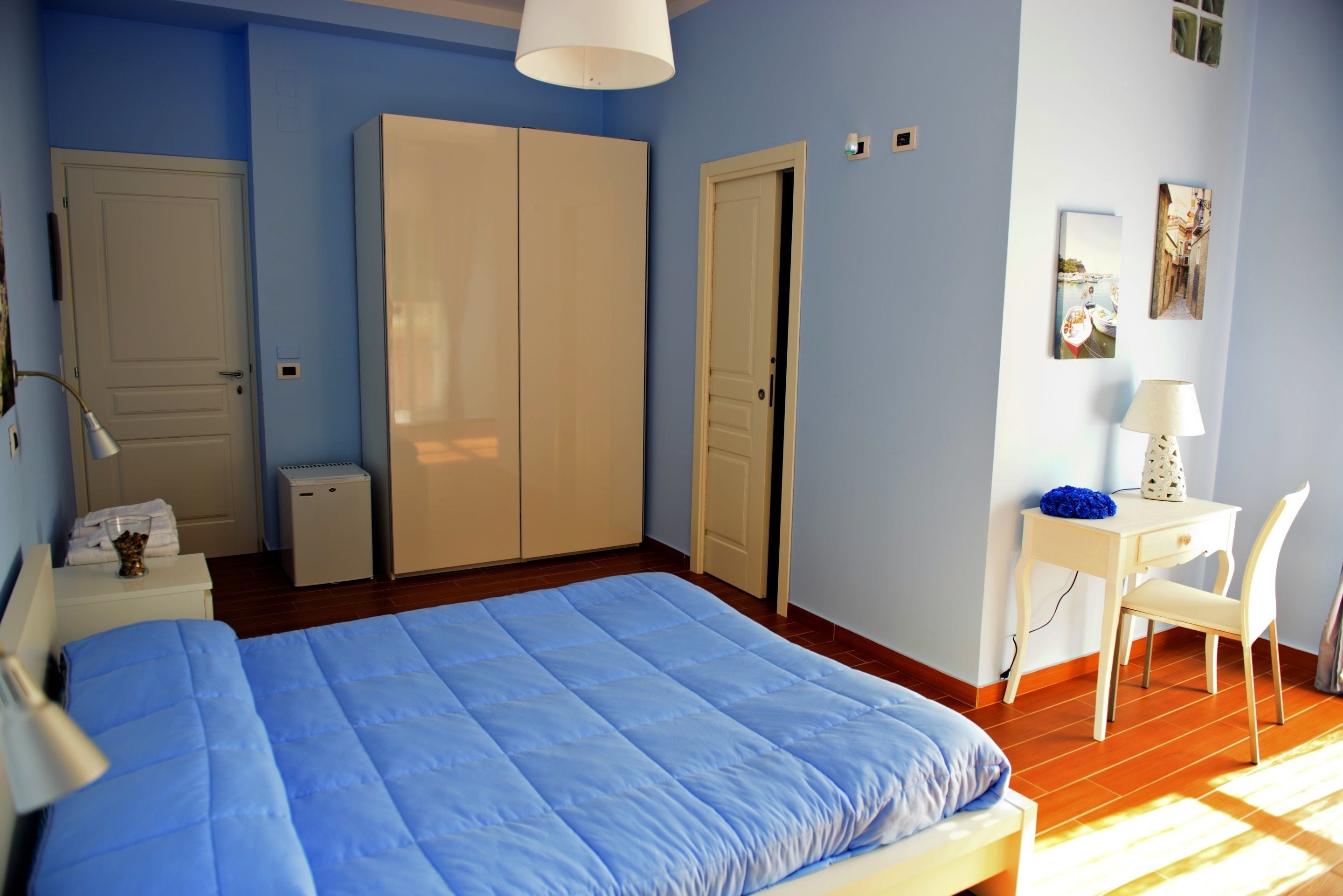 A bedroom with a large bed, a wardrobe, a desk with a chair, and a small table with a lamp.
