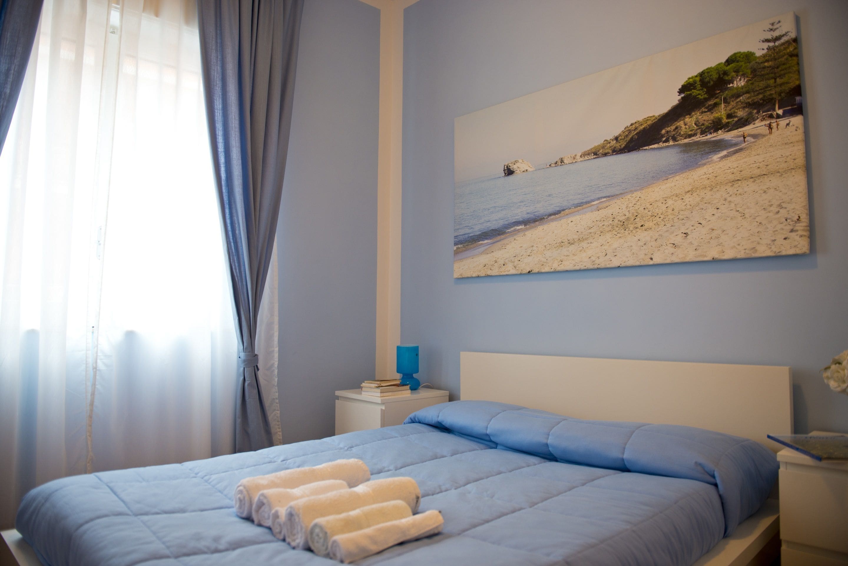 Une chambre à coucher avec un lit, une table de chevet, un tableau représentant une plage et une fenêtre avec des rideaux.