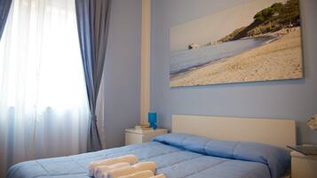 Une chambre à coucher avec un lit, une table de chevet, un tableau représentant une plage et une fenêtre avec des rideaux.