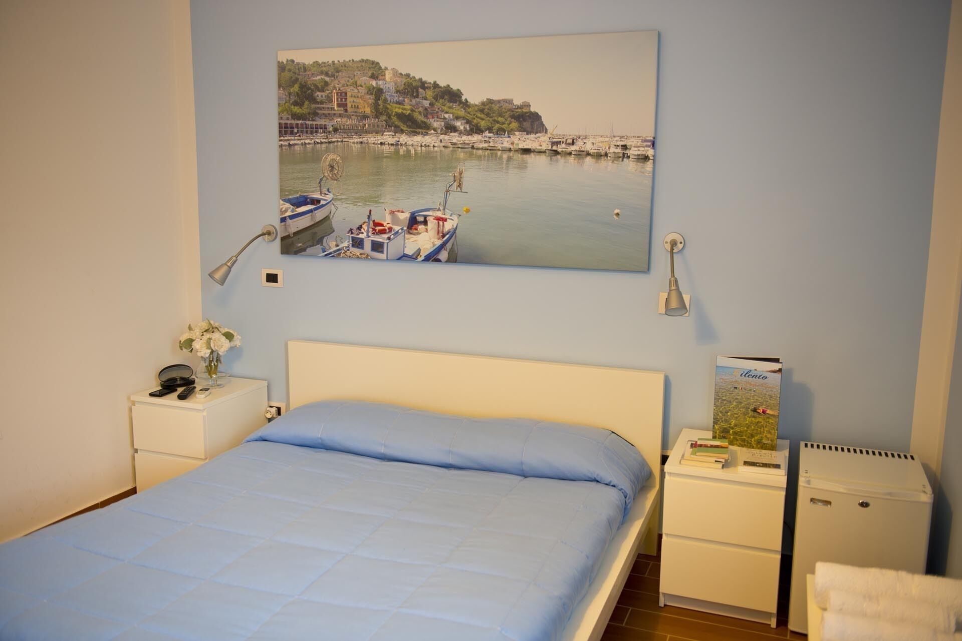 Un dormitorio con cama, mesita de noche, lámpara, pequeña mesa con un jarrón de flores, cuadro colgado en la pared que muestra una escena costera y un refrigerador.