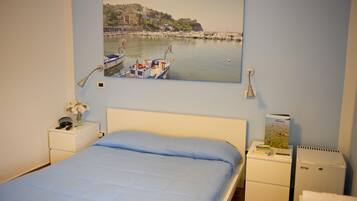 Un dormitorio con cama, mesita de noche, lámpara, pequeña mesa con un jarrón de flores, cuadro colgado en la pared que muestra una escena costera y un refrigerador.