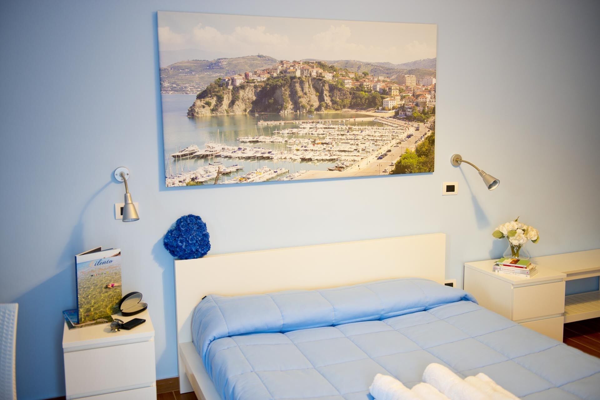 A bedroom with a bed, a nightstand, a lamp, a wall-mounted picture of a coastal town, and a vase with flowers.