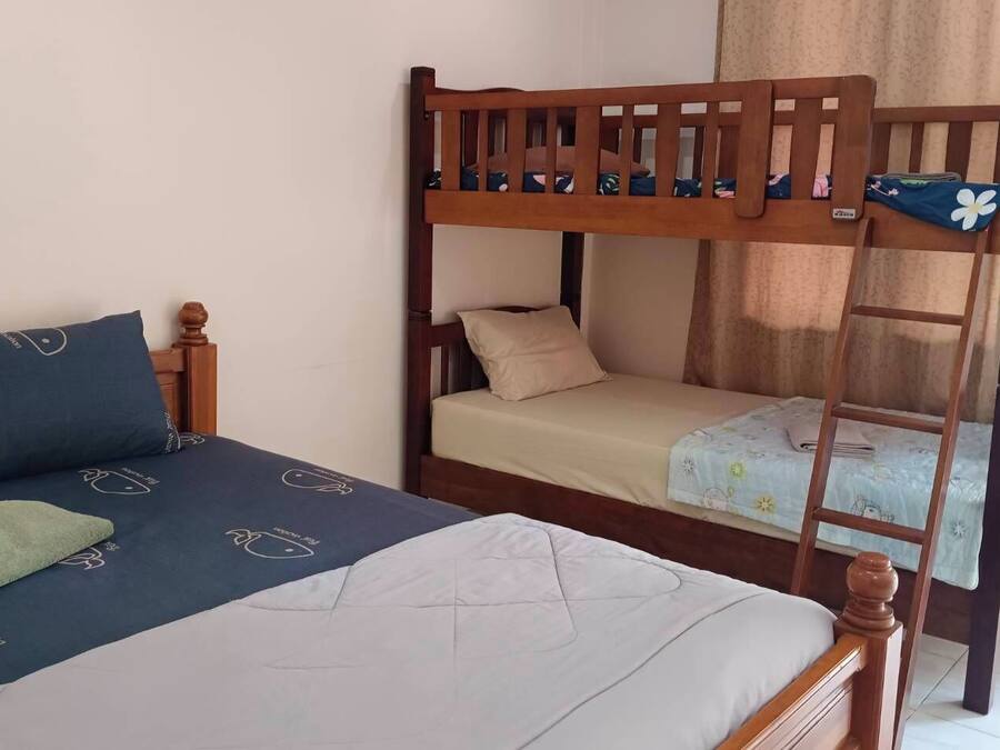 A room with a bunk bed and a single bed. The bunk bed has a wooden frame and a mattress with a floral pattern. The single bed has a white mattress and a blue bedspread with a white pattern. There is a green towel on the single bed.