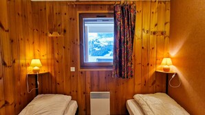 Une pièce aux murs en bois, avec deux lits, une fenêtre avec des rideaux et des lampes fixées au mur.