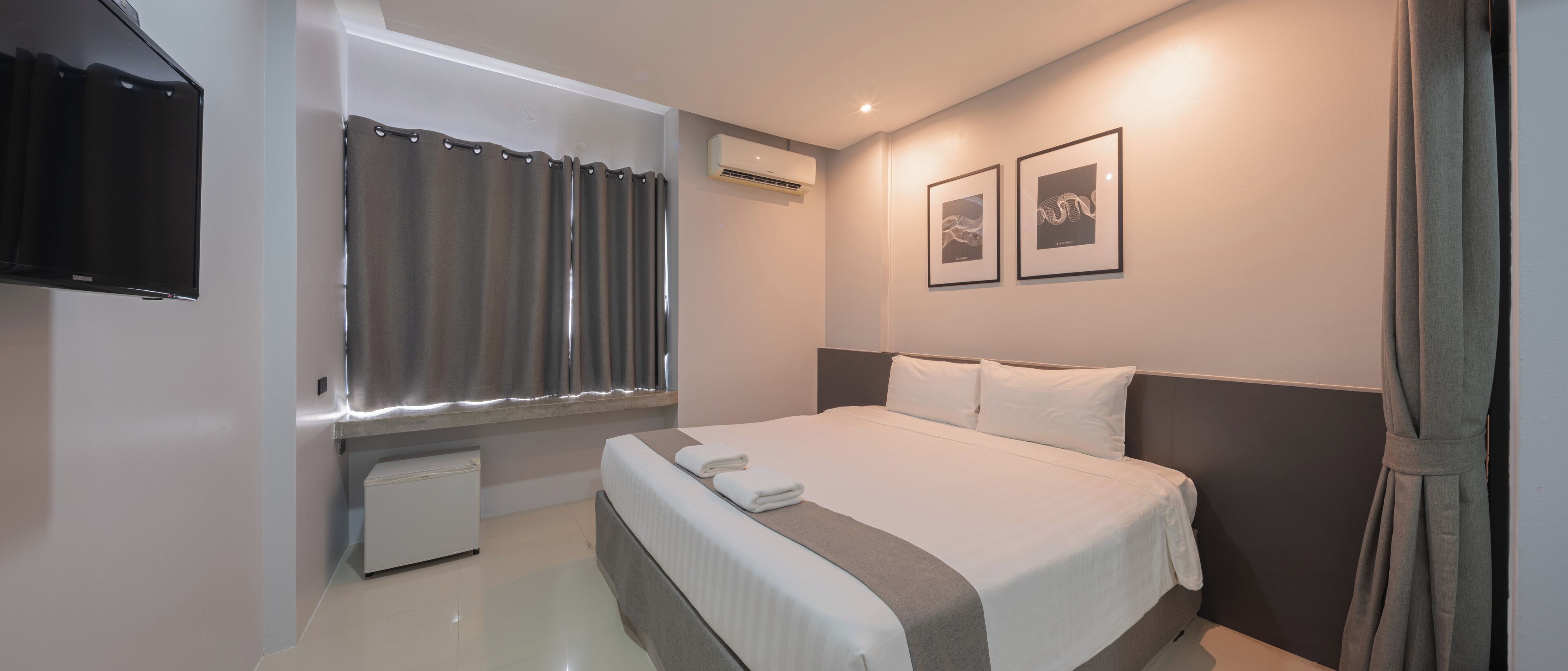 Una habitación de hotel con cama, televisión de pantalla plana, aire acondicionado, un refrigerador pequeño y dos cuadros en la pared.