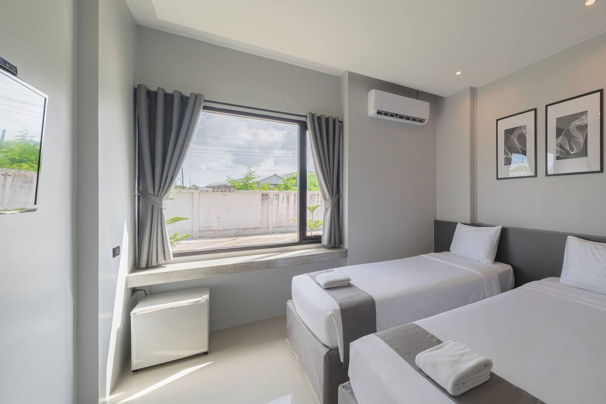 A hotel room with two beds, a window with curtains, an air conditioning unit, and two framed pictures on the wall.