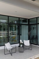Un área de estar exterior moderna con dos sillas, una mesita pequeña, ventanas de cristal amplias y vista a la calle.