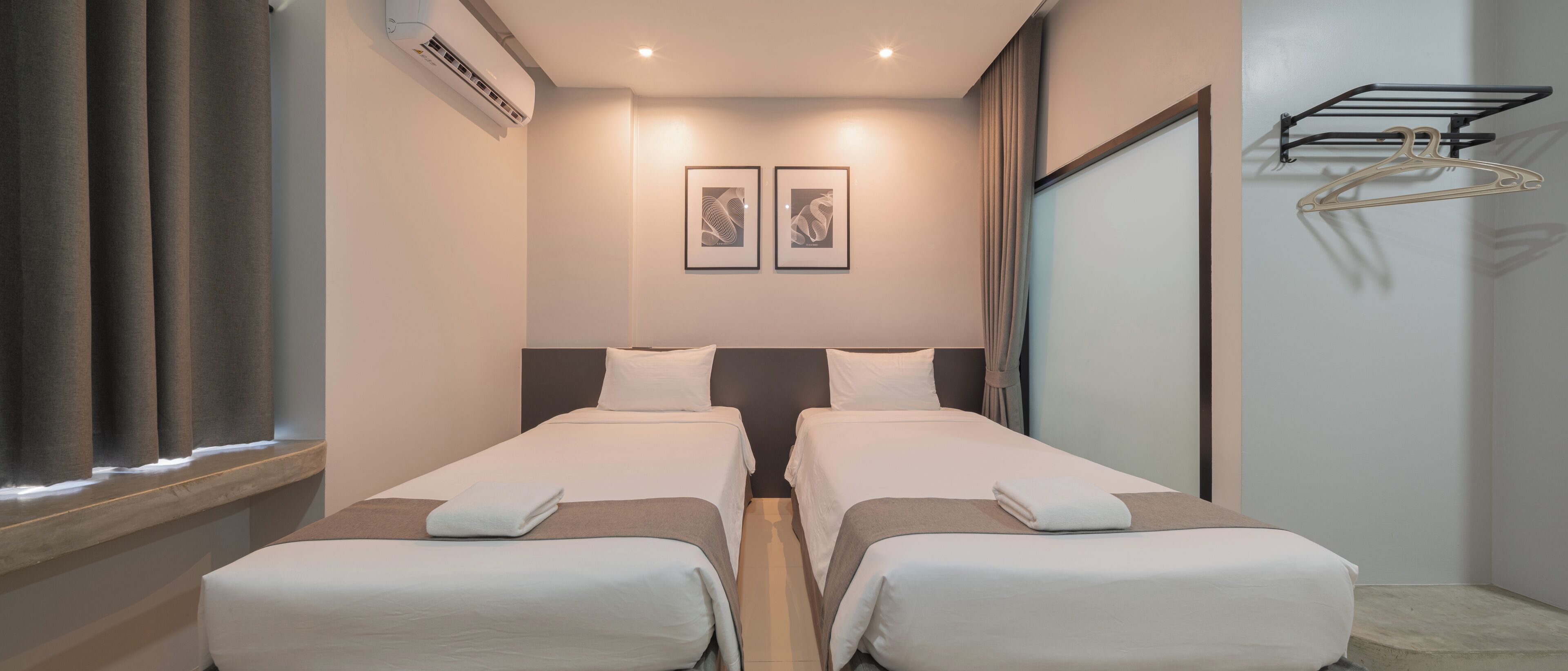 A hotel room with two beds, a window with curtains, an air conditioning unit, and a wall-mounted drying rack.
