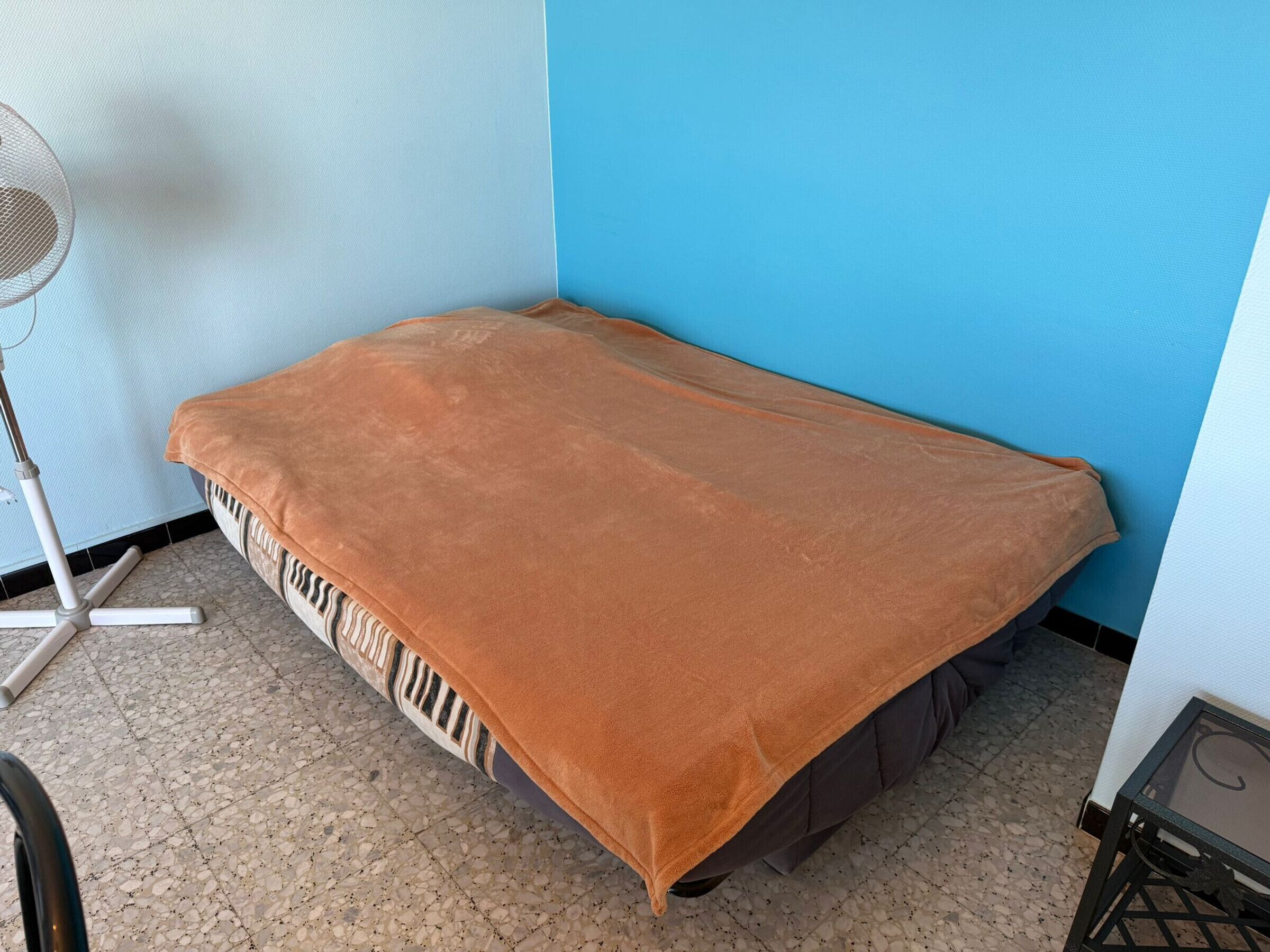 Une petite chambre avec un lit simple, une couverture marron, un ventilateur sur pied et un sol carrelé.