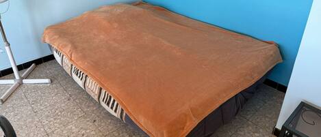 Une petite chambre avec un lit simple, une couverture marron, un ventilateur sur pied et un sol carrelé.