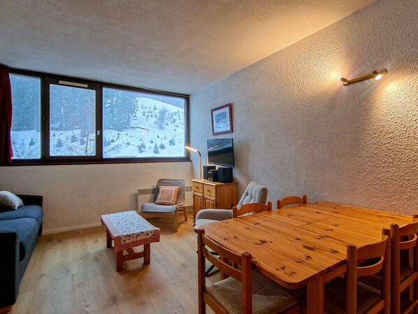 A cozy room with a wooden dining table, two chairs, a small table with a patterned tablecloth, a sofa, a TV, and a window with a snowy view.