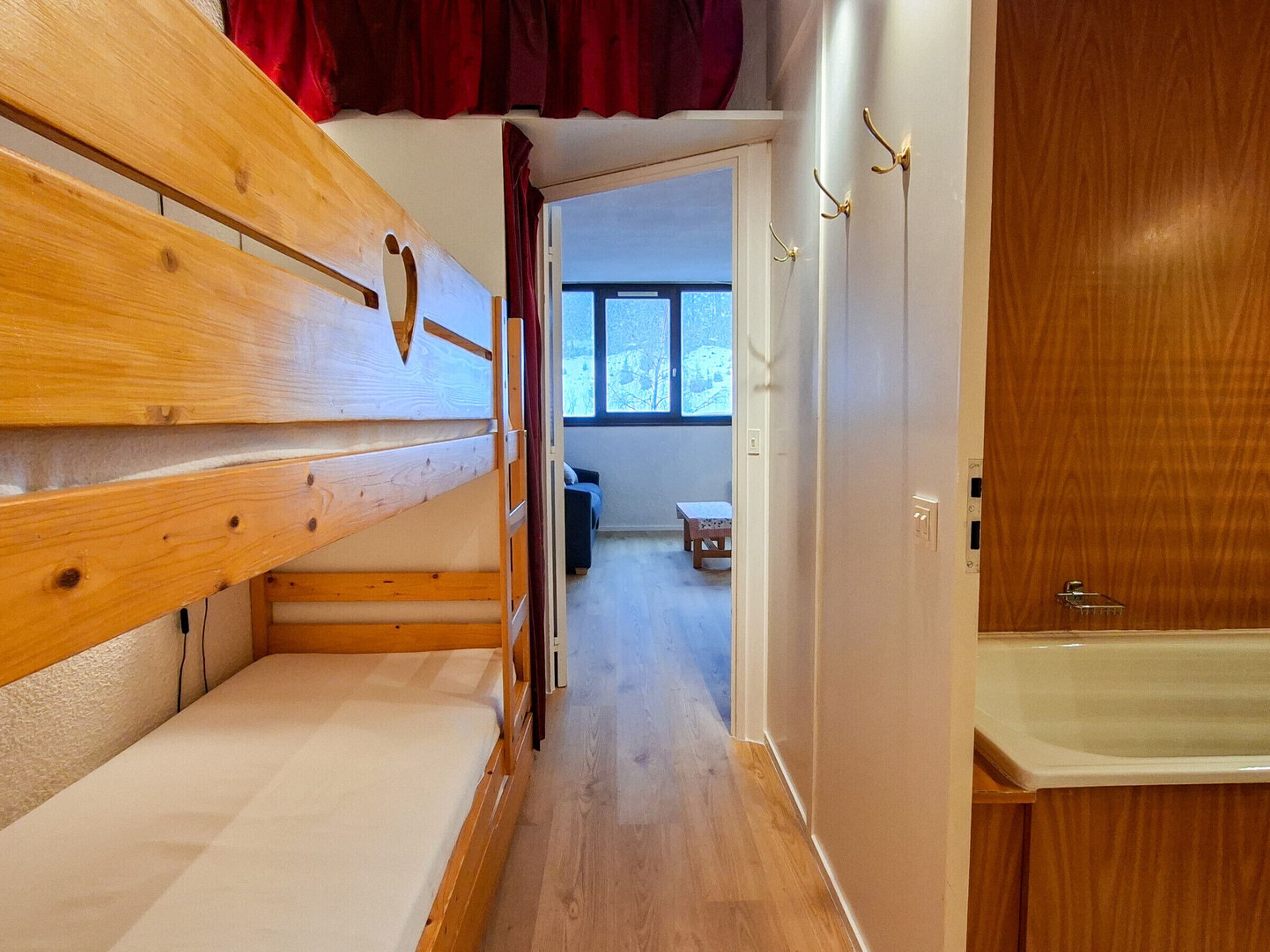 Une pièce avec un lit superposé en bois, une petite table, un banc et, par une porte ouverte, on aperçoit une autre pièce avec une fenêtre.
