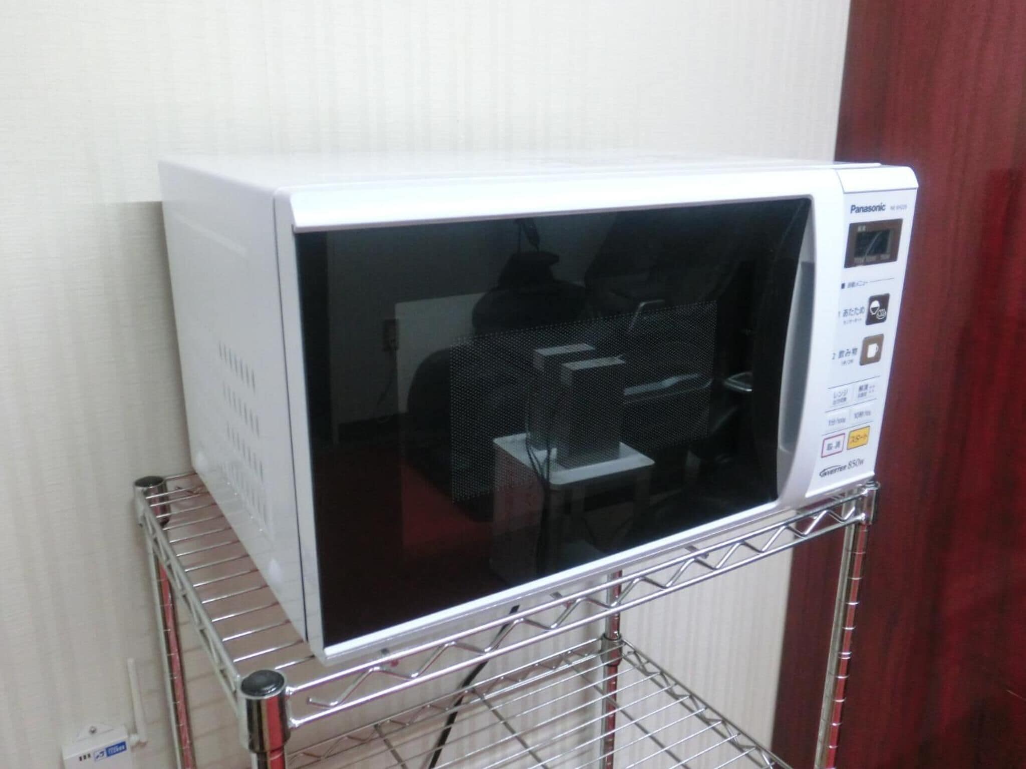 A white microwave oven on a metal shelf in a room with a red curtain and a white wall.