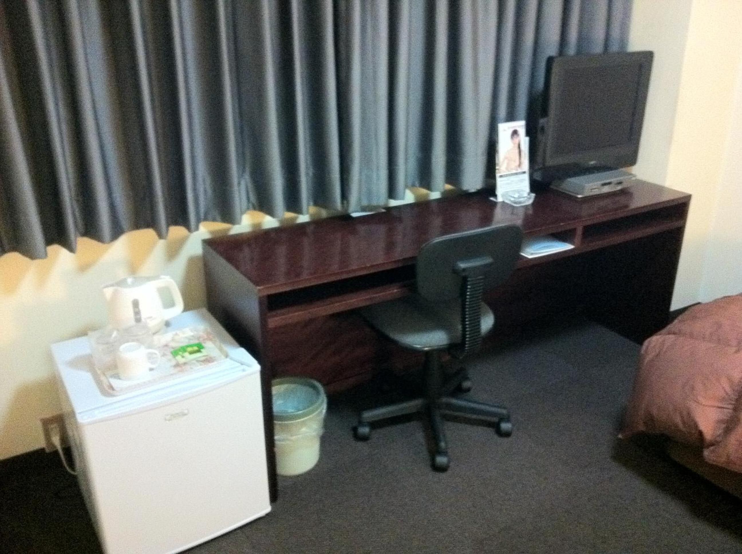 A hotel room with a wooden desk, a black office chair, a television, a mini-fridge, and a bed with a pink blanket.
