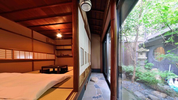 A traditional Japanese room with tatami flooring, a low wooden bed, and a view of a garden through a large window. - Takeya Kitano Honkan (Kyoto)