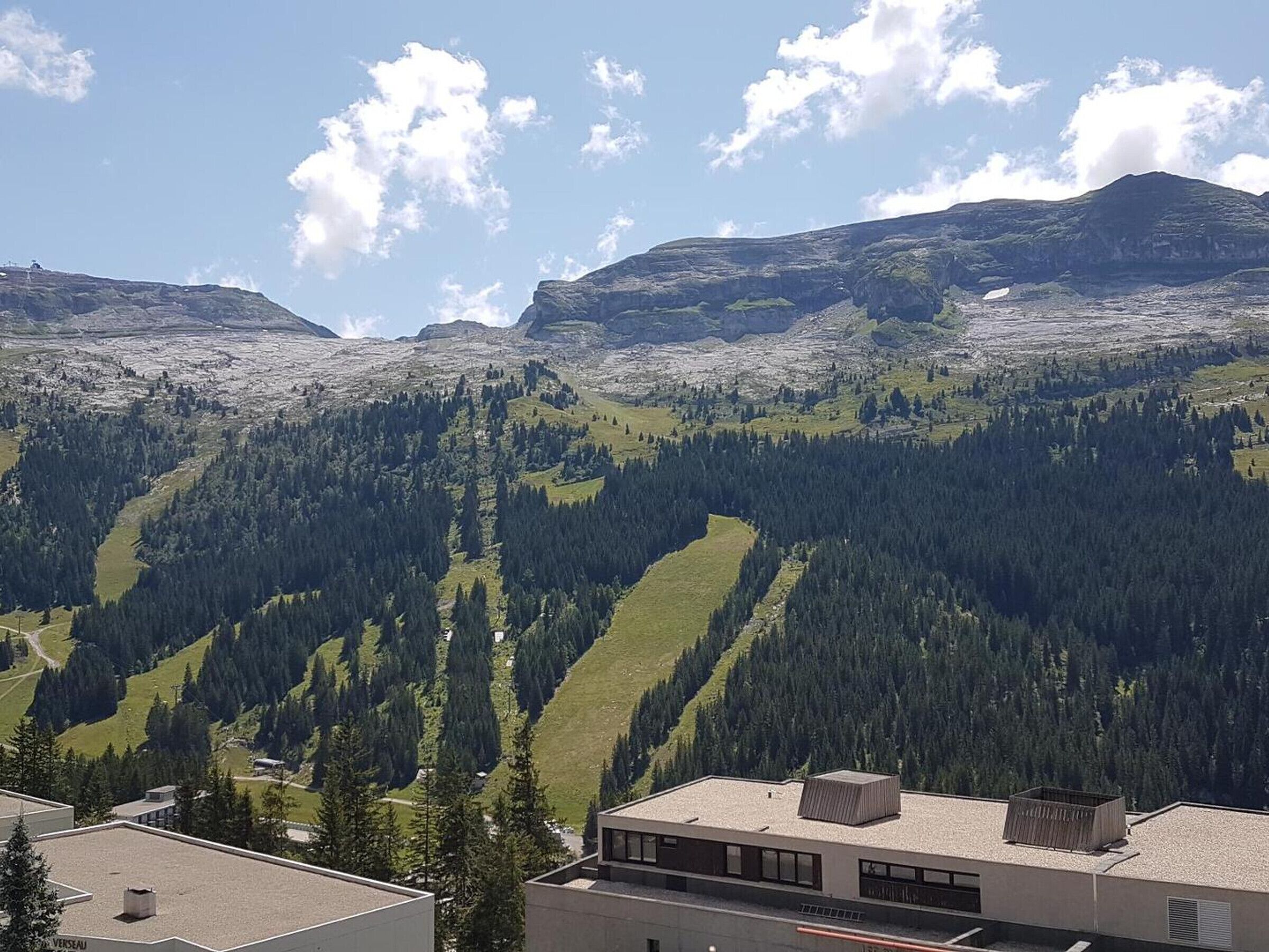 Ein modernes Gebäude mit Flachdach und erkennbarem Schornstein, eingebettet in eine gebirgige Landschaft mit dichten Wäldern und blauem Himmel.
