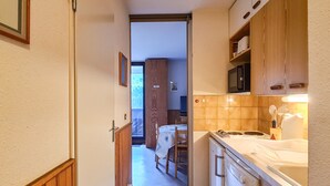 A narrow kitchen with wooden cabinets, a microwave, and a sink. A dining area is visible in the background. - Bright studio in Flaine center with balcony, sleeps 4 (Flaine)