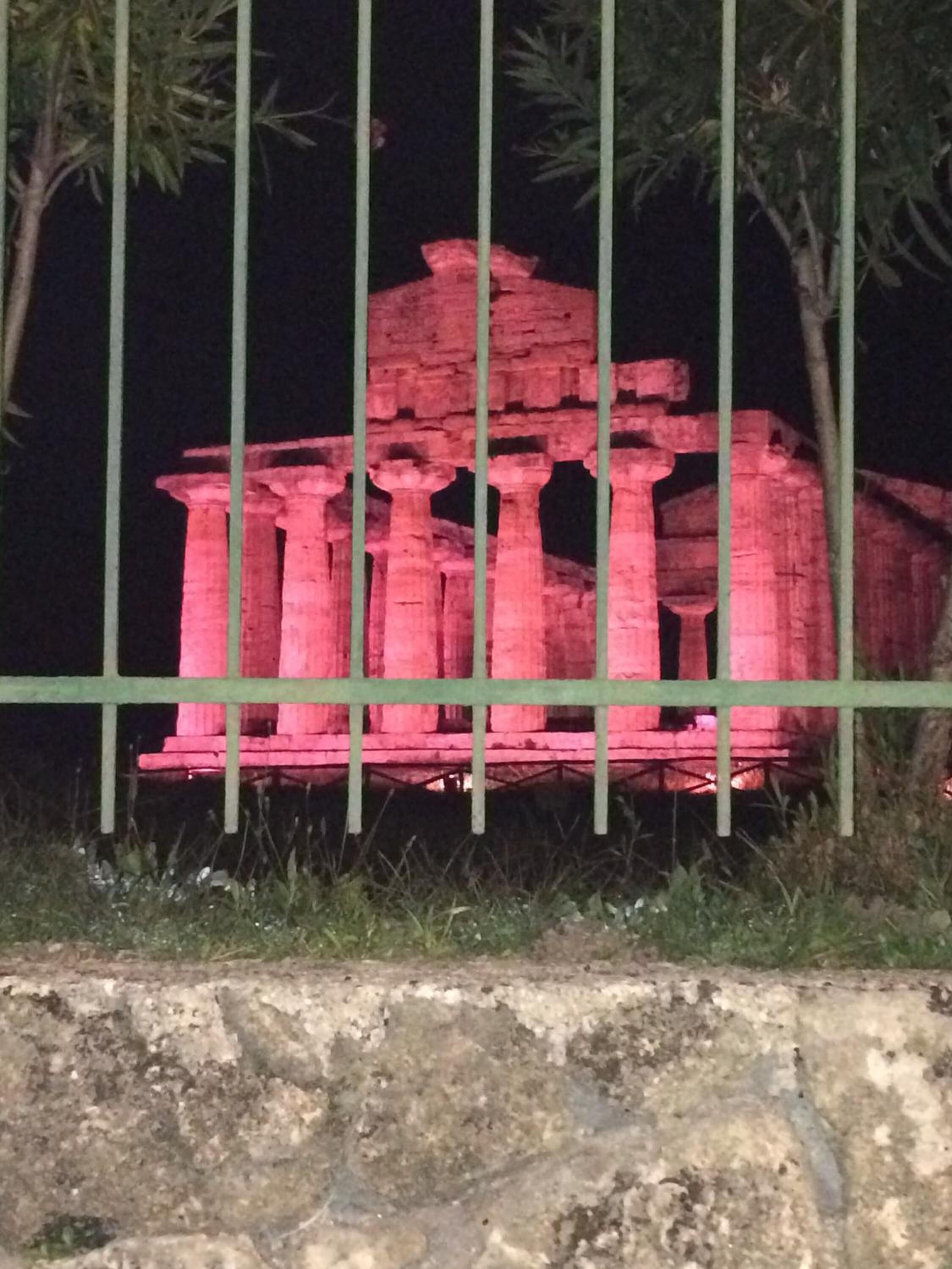 A stone structure illuminated in red, surrounded by a metal fence and greenery.
