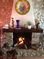 Una chimenea de piedra con fuego encendido, un plato decorativo encima y varios objetos en la repisa, incluyendo un florero, una planta y una botella.