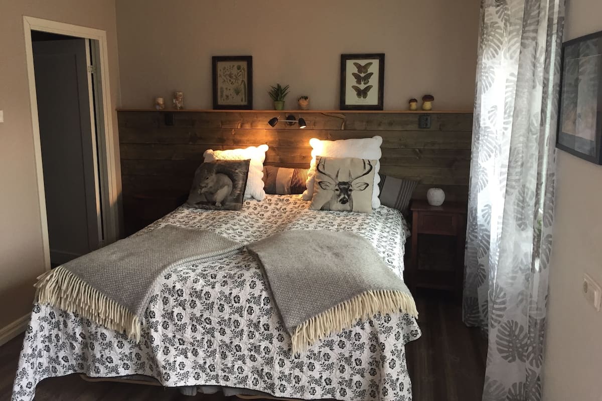 Une chambre à coucher comprenant un lit, deux oreillers, une table de chevet, une commode, une porte et un mur orné de tableaux.