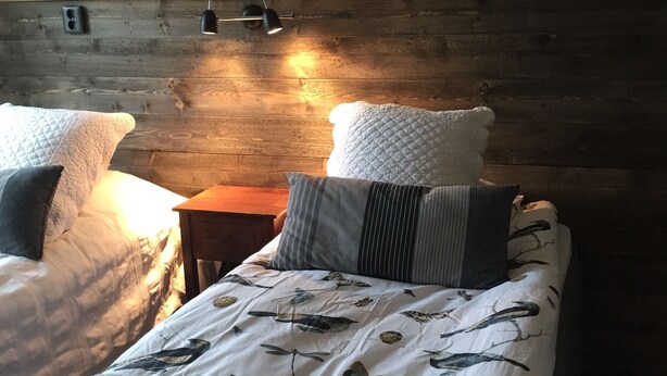 A cozy bedroom with two beds, a wooden headboard, and a wall-mounted shelf with decorative items. - KUSKAHUSEN VANDRARHEM (Brosarp)