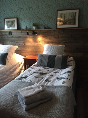 A cozy bedroom with two beds, a wooden headboard, and a wall-mounted shelf with decorative items. - KUSKAHUSEN VANDRARHEM (Brosarp)