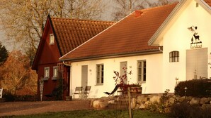 A two-story house with a red roof and white walls, featuring a chimney, a front porch with chairs, and a sign with a deer and trees. - KUSKAHUSEN VANDRARHEM (Brosarp)