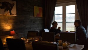 A dimly lit room with a wooden wall, a table with chairs, and a window with curtains. - KUSKAHUSEN VANDRARHEM (Brosarp)
