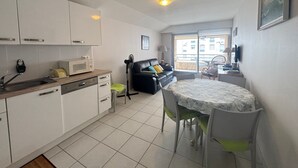 A compact living space with a kitchenette, a dining area with a table and chairs, a black leather sofa, and a view through a large sliding glass door to an outdoor balcony. - Downtown Apartment Steps from Beach with Garage (Les Sables-d'Olonne)