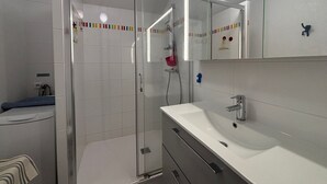 A modern bathroom with a glass shower enclosure, a white sink, and a mirror with a colorful border. - Downtown Apartment Steps from Beach with Garage (Les Sables-d'Olonne)