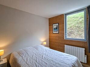 Une chambre à coucher avec un lit, une fenêtre donnant sur un paysage verdoyant, un tableau accroché au mur et un radiateur.