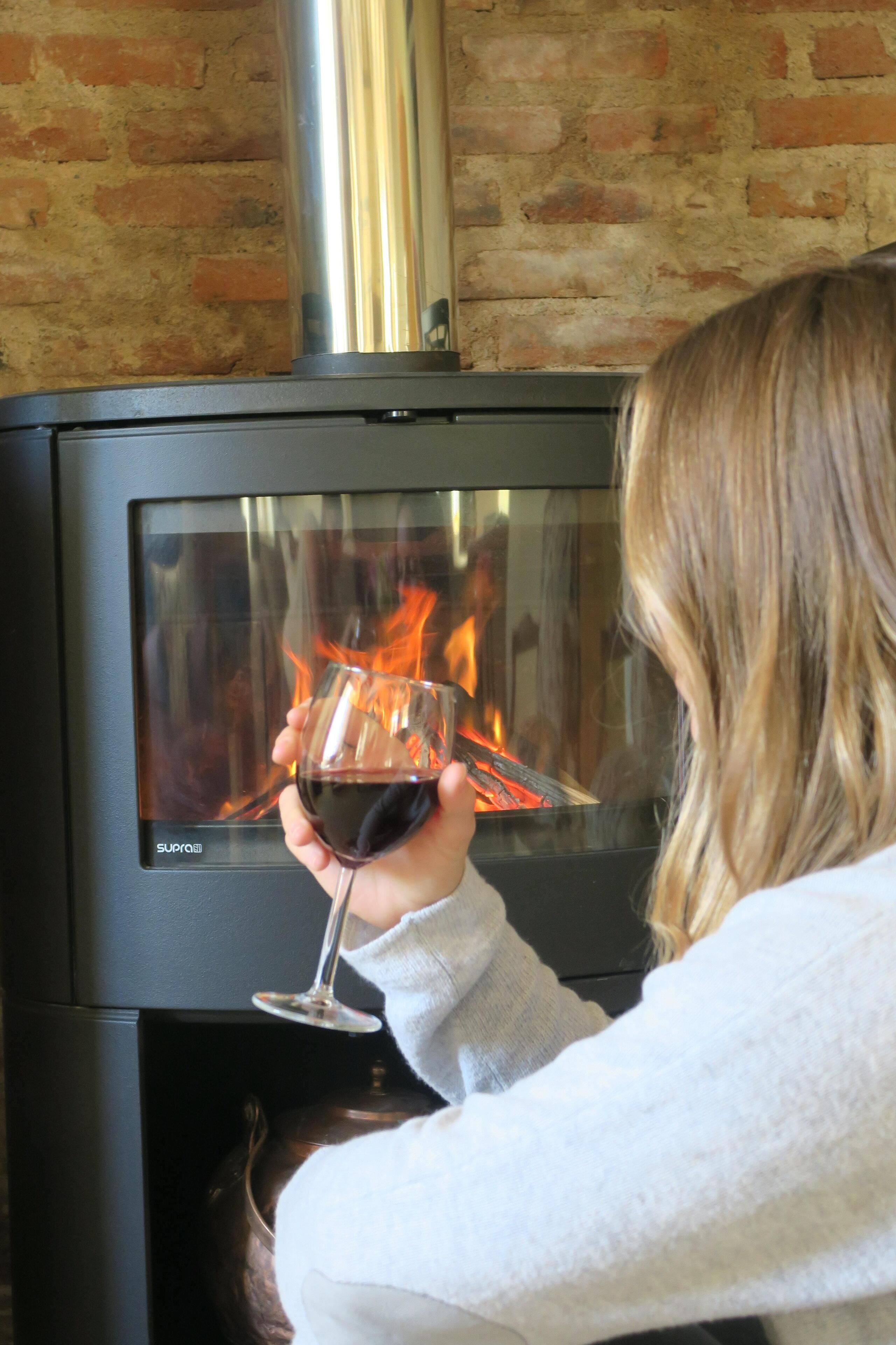 Een persoon houdt een glas rode wijn vast voor een moderne haard met een glazen deur.