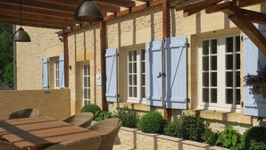 An outdoor patio with a wooden table and wicker chairs, stone walls, and blue shutters on windows. - HOUSE OF CEDAR, BEAUTIFUL STONE HOUSE IN DORDOGNE. (Auriac-du-Périgord)