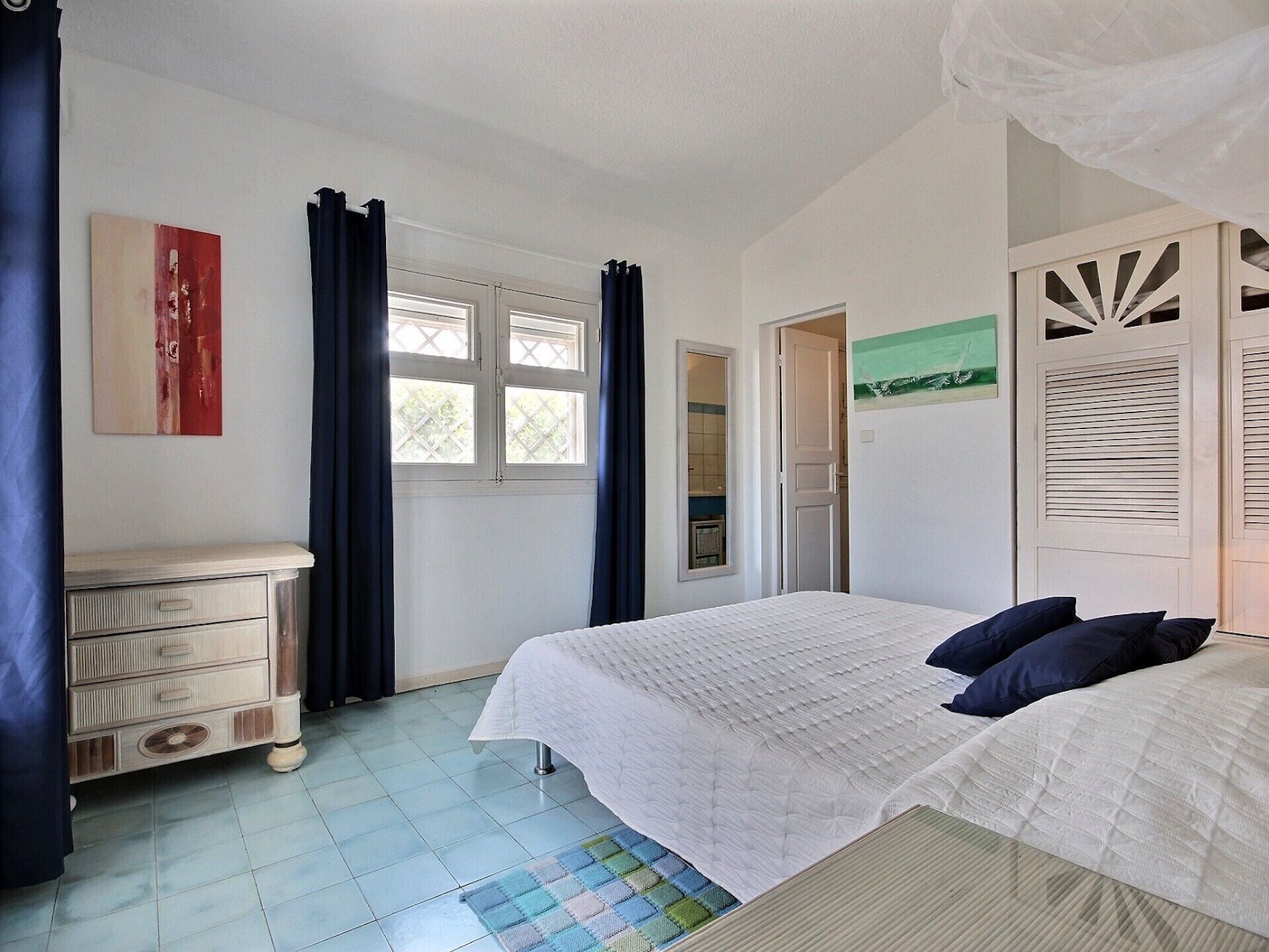 Ein Schlafzimmer mit zwei Betten, einem Schminktisch, einem Fenster mit Vorhängen und einer Tür zu einem anderen Raum.
