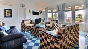 Ein Wohnzimmer mit Schachbrettmuster-Boden, Korbmöbeln, einem Flachbildfernseher und großen Fenstern mit Vorhängen.