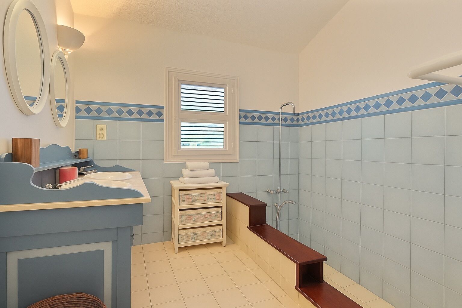 Un baño con pared de azulejos azules y blancos, un mueble de tocador de madera con espejo, una ventana con persianas y una zona de ducha.