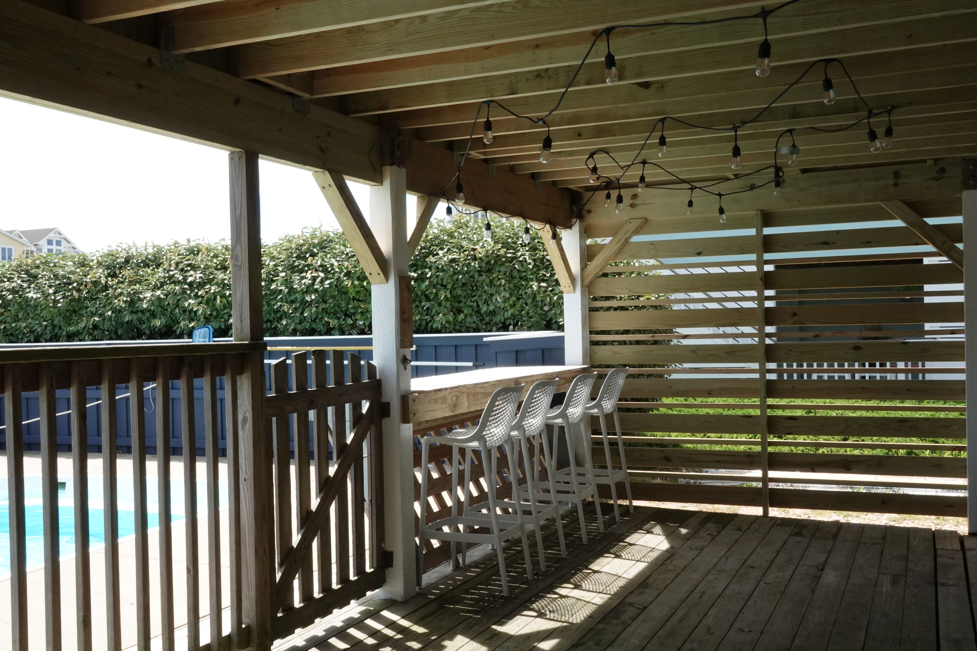 A covered outdoor area with wooden beams and string lights, featuring a pool and a wooden fence.