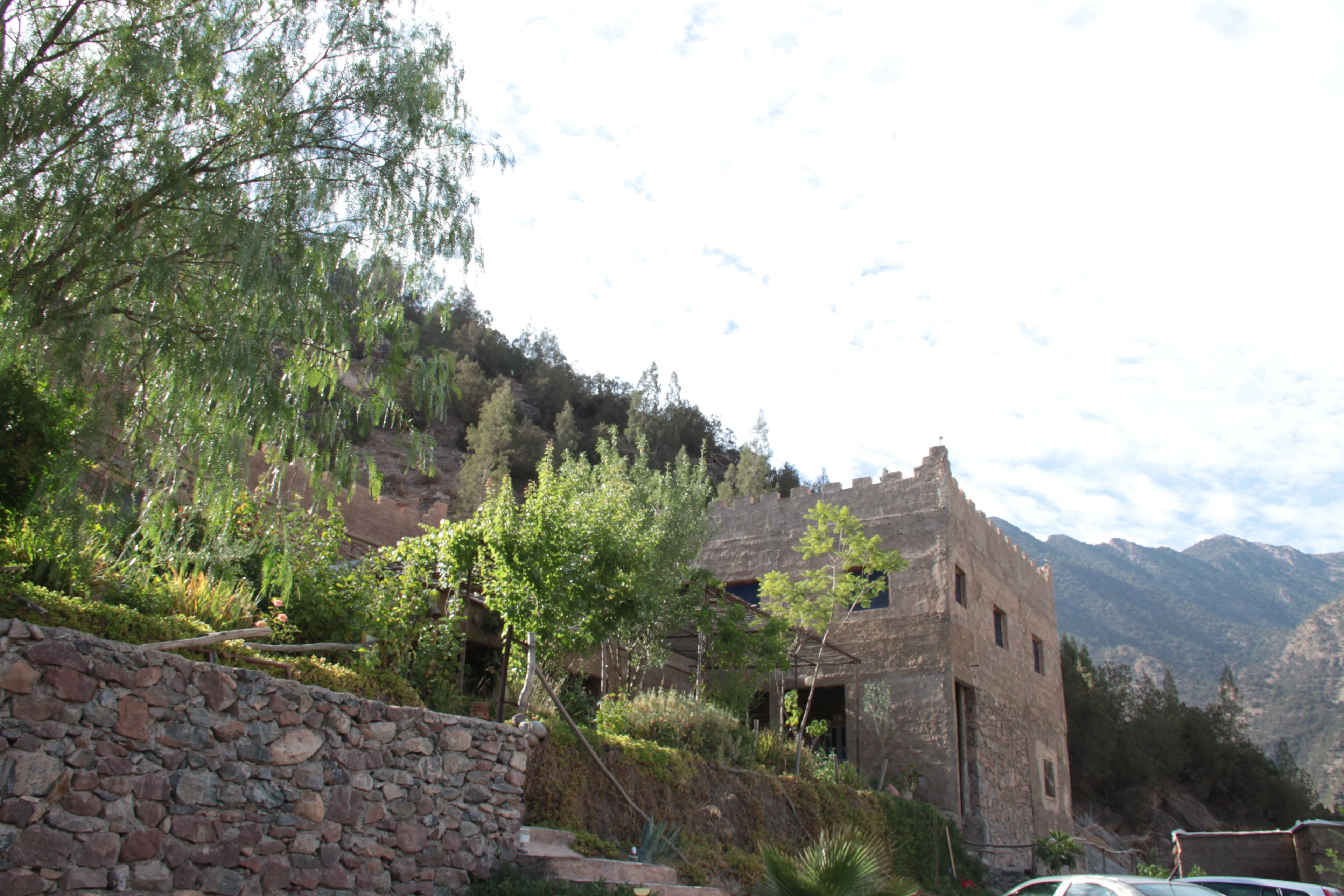 Un edificio de piedra con arquitectura rústica de estilo medieval, rodeado de abundante vegetación y montañas al fondo.