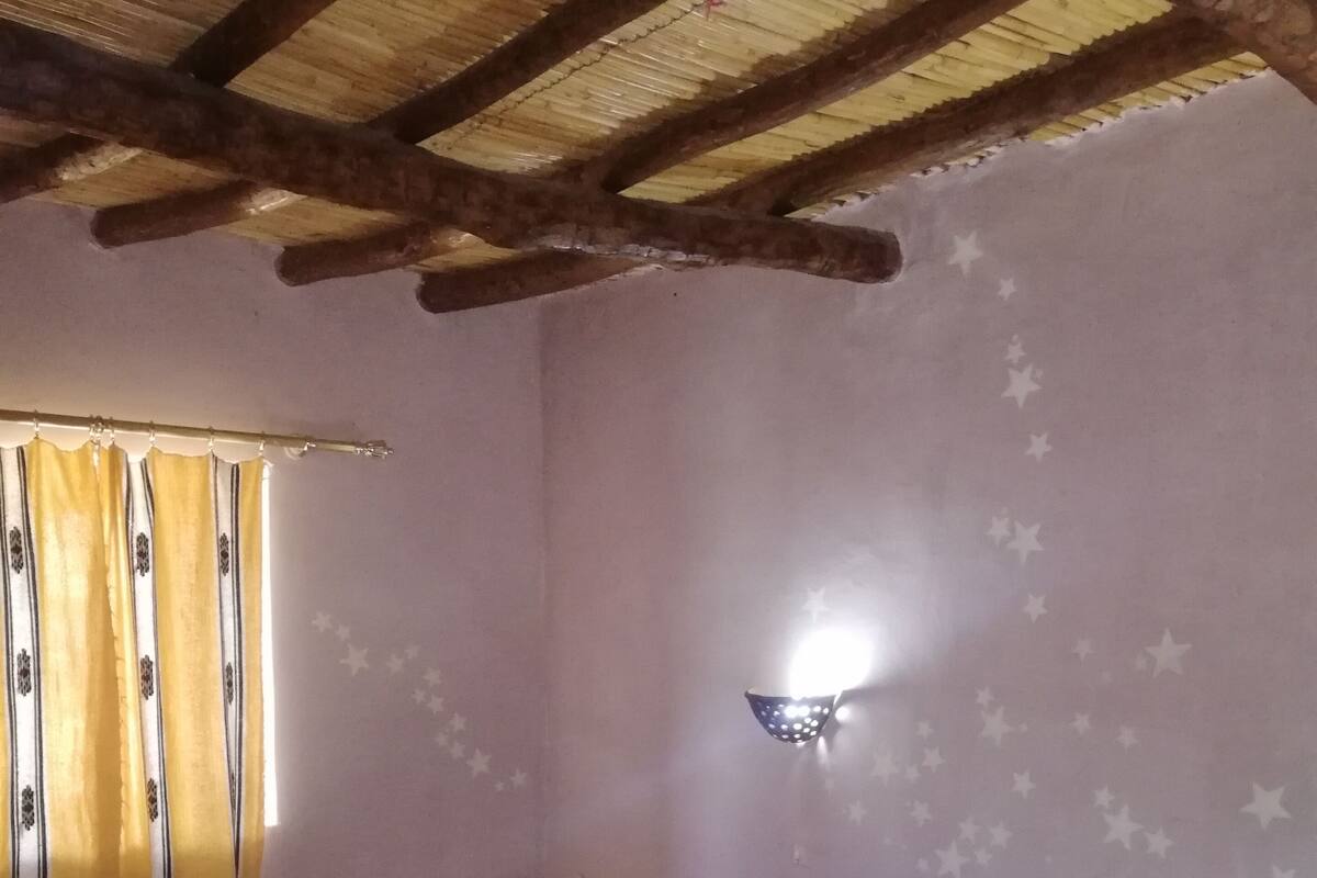 Makuuhuone, jossa on puinen katto, kuviollinen päiväpeite sängyllä, yöpöytä ja seinään kiinnitetty valaisin.